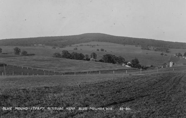 View of farmland near Blue Mounds, with possibly property markers horizontal, across the center, and some distant farm houses and a barn. Caption reads: "Blue Mound-1756 ft. Altitude. Near Blue Mounds, Wis."
