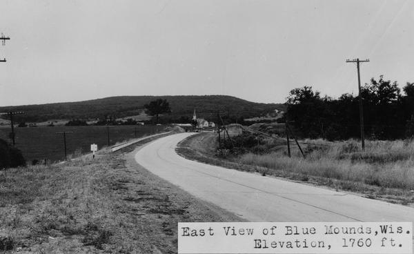 View of rural Blue Mounds, with a two-lane, paved road. The caption at the bottom reads: "East View of Blue Mounds, Wis. Elevation, 1760 ft.".