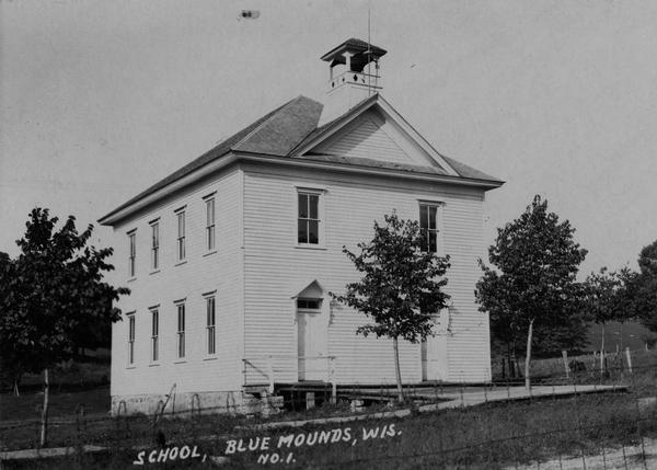 Spring or summer view of a large, white school house, and a few trees in front of it. Caption reads: "School, Blue Mounds, Wis."