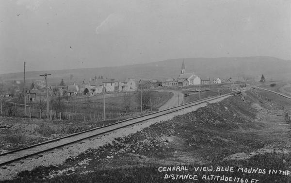 Distant view of the town, with railroad tracks in foreground. The church is the highest point in the image. "General View. Blue Mounds in the Distance. Altitude 1760 ft."