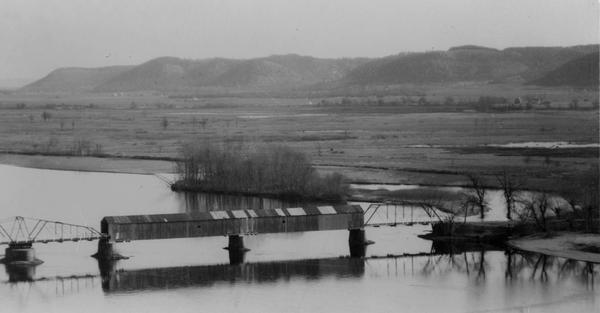 Elevated view of a covered railroad bridge over a calm body of water, with bluffs in the background.