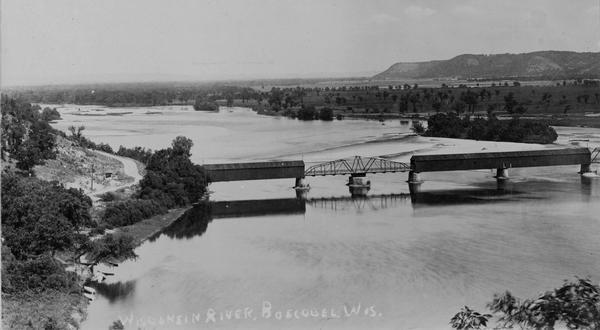 Elevated view of railroad bridge over the river. Part of the bridge is covered. Bluffs are in the background.