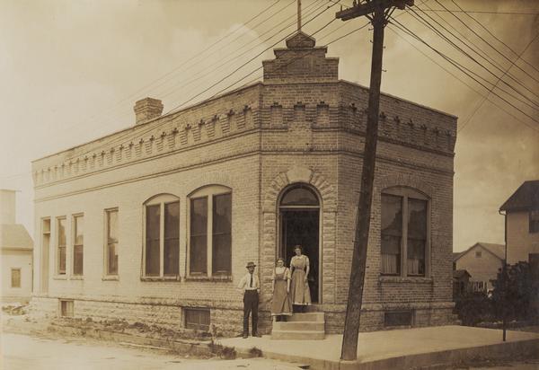 View from street towards the Brillion News building. A man and two women are posed in the doorway at the corner of the building.