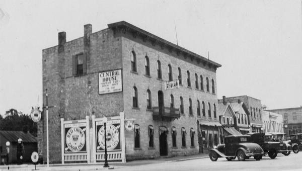 Exterior view from street of Central House, with advertisements on the side of the building for Red Crown Gasoline. The Order of the Gideons was founded here. Cars are parked outside the hotel.