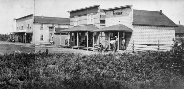 View of three buildings; a store, hotel and post office. A group of men are gathered on the porch at the post office.