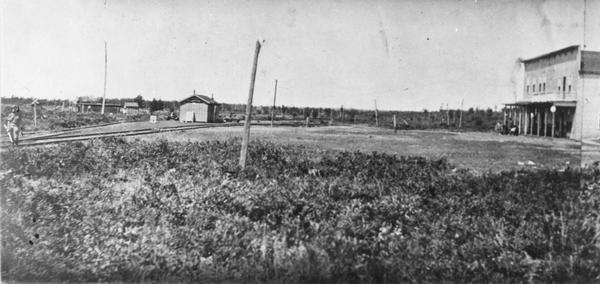 View looking towards railroad tracks and a station on the left, with a store and hotel on the right. A man is standing near the railroad tracks on the far left.