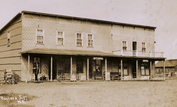 Storefront with goods on the left side of the building, and on the front porch. The sign on the left reads: "Store", and the sign on the right reads: "Hotel". Caption reads: "Boulder Jct., Wis."