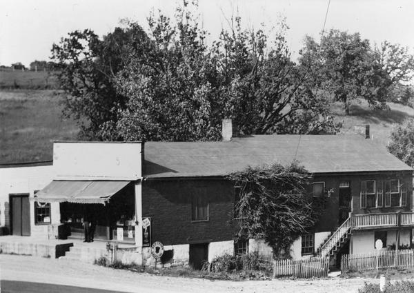 Ivey and Webb general store, with trees and a hill behind, and in front a man standing under the store's awning.