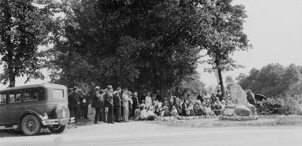 View of crowd of people under the trees near the trading post marker in Butte des Morts.