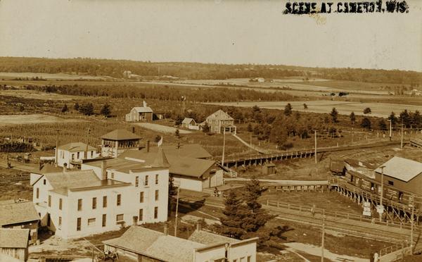Aerial view of town, with several large buildings along railroad tracks in the foreground, and fields and trees in the distance. Pedestrians are walking along elevated boardwalks, and railroad cars are on curved railroad tracks near a building on the right.