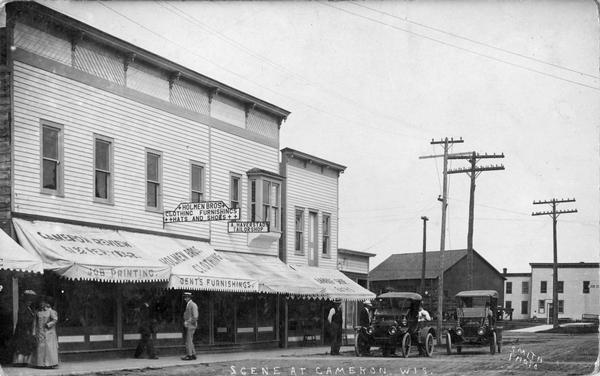 Downtown street in Cameron, with buildings with awnings over the show windows, including Holmen Bros. Clothing store, Gent's Furnishings, and A. Haverstad's Tailorshop. Pedestrians are on the sidewalk, and two automobiles are parked on the unpaved street.