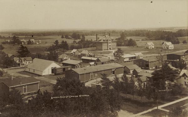 Caption reads: "Birdseye [<i>sic</i>] View of Cameron Wisconsin." Railroad cars are on railroad tracks between buildings, and houses and what may be the high school are in the background beyond.