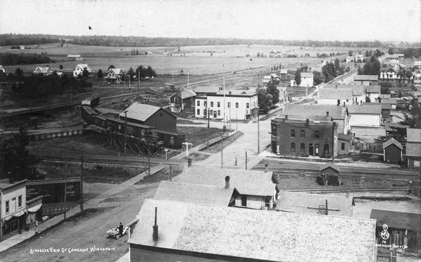 Elevated view of Cameron. Rooftops are in the foreground, and an unpaved street is along the left. Railroad tracks cut across the street, with railroad cars just behind on elevated tracks near buildings. In the distance are houses, fields, and tree-lined hills.
