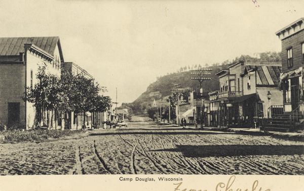 Unpaved road in downtown Camp Douglas. There is a bluff in the background on the right. Caption reads: "Camp Douglas, Wisconsin."