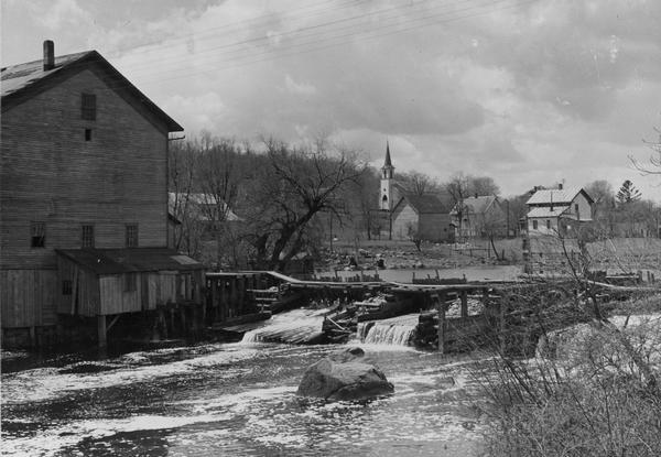 Elevated view from shoreline towards the opposite shoreline towards the Buettner mill on the left, and the river with a dam, on the right. In the background is a church building and other buildings.