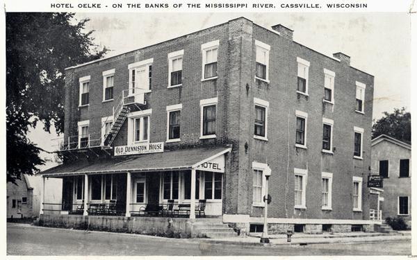 Caption reads: "Hotel Oelke - on the banks of the Mississippi River, Cassville, Wisconsin." Sign on the hotel reads: "Old Denniston House".  This view shows the hotel after the removal of the 4th floor during the 1940s.