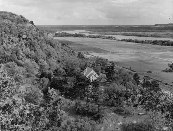 Elevated view from bluff of a house in Cassville surrounded by bluffs, trees and farmland. Railroad tracks run between the crops and the trees, and the Mississippi River is in the background.