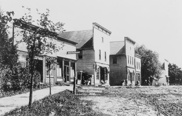 View of storefronts, a dirt road, and a plank sidewalk. One of the storefronts is a restaurant. Caption reads: "Cazenovia, Wis."