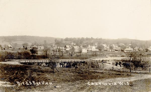 Bird's-eye view of Cazenovia, with a bridge in the foreground, the town,  and in the distance a low hill in the background. Caption reads: "Bird's Eye View, Cazenovia Wis."