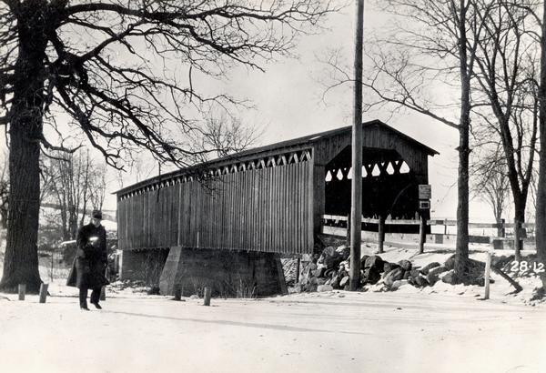 Southwest view of a covered bridge over Cedar Creek, with a man walking in the snow nearby.