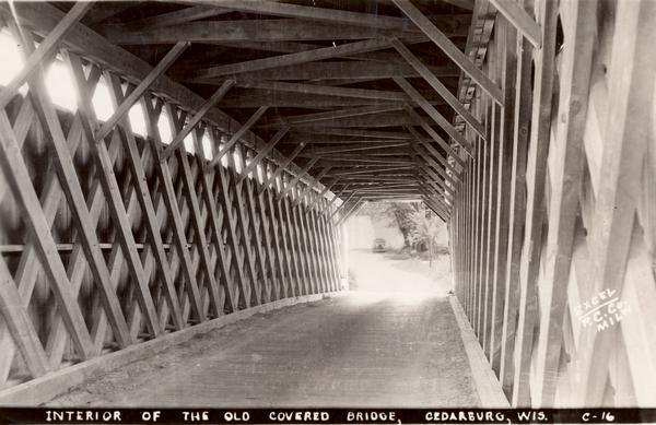 Last covered bridge to remain standing in Wisconsin. In the distance an automobile is parked on the side of the road in the shade of a tree. Caption along bottom reads: "Interior of the Old Covered Bridge, Cedarburg, Wis."