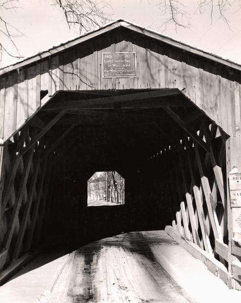 View through the covered bridge over Cedar Creek in Ozaukee County on highway, about one mile north of Cedarburg. The last remaining covered bridge in Wisconsin. Snow is on the ground. A plaque above the bridge reads: "1876 1955 Last Covered Bridge in Wisconsin — Placed By — Port Washington Chapter of the Daughters Of The American Revolution".