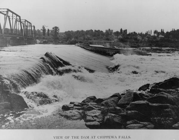 Dam and bridge near Chippewa Falls, looking across the river towards a few buildings on the opposite bank. Caption at bottom reads: "View of the Dam at Chippewa Falls."