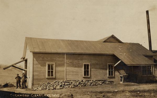 View of the side of the creamery, with two men standing at an open door at the end of the building on the left. Caption reads: "Creamery Clear Lake Wis."