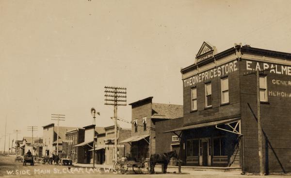 West side of Main Street, with "The One Price Store" in the right foreground, and other commercial buildings further down the street. There are large utility poles along the sidewalk. In the street are horses and horse-drawn wagons. A windmill is in the background. Caption reads: "W. Side Main St. Clear Lake, Wis."