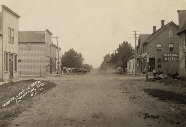 Main Street looking north. "Hotel and Saloon" are on the right side, along with other buildings down the right and left side. Main Street is a dirt road. There are some children standing on a corner, and two horses and horse-drawn wagons in the distance.