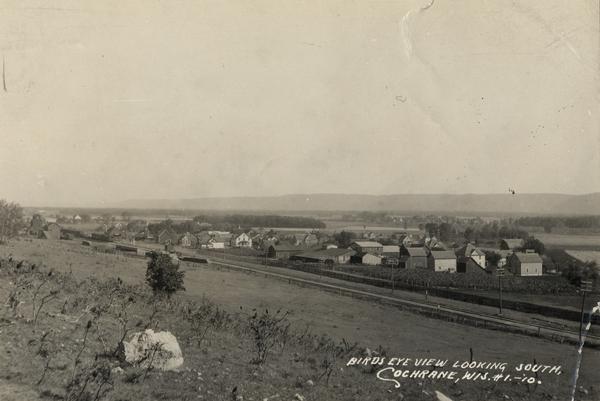Slightly elevated view of Cochrane from a field. A road or highway stands between the field and Cochrane. Caption reads: "Birds [<i>sic</i>] Eye View Looking South."