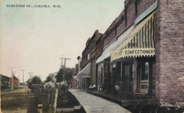 View down sidewalk on right side of the business district. There is an awning that reads "Restaurant" and "Fine Confectionary". Horses and a horse-drawn wagon are on the left along the curb. Caption reads: "Business St., Coloma, Wis."
