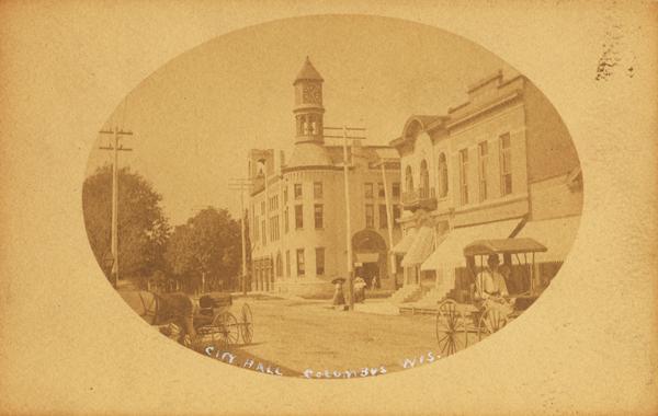 View down street towards City Hall, and some nearby buildings. Two horse-drawn wagons are on the street, along with a horse and riders.  There are pedestrians in front of City Hall, including a woman with a parasol.