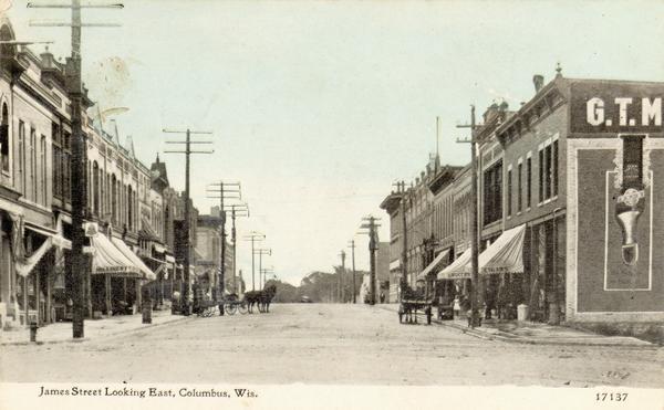 View down center of James Street with a couple of horse-drawn wagons along the curbs. Storefronts line both sides of the street. Caption reads: "James Street Looking East, Columbus, Wis."
