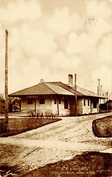 View down unpaved road towards the railroad station. Caption reads: "R. R. Station, Columbus, Wis."
