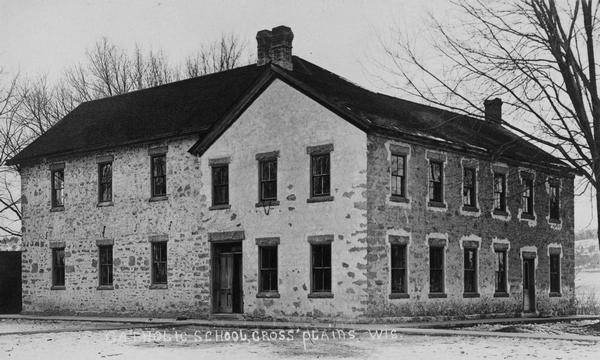 View of the exterior of a Roman Catholic school built of stone with many windows. Sidewalks run along two sides. Trees can be seen behind and on the right. Text reads: "Catholic School, Cross Plains, Wis."