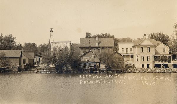 View of several Cross Plains buildings across a millpond. There is a bell mounted on a metal tower near the shoreline. Caption reads: "Looking North From Mill Pond, Cross Plain, Wis."
