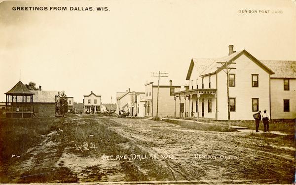 View down unpaved street towards commercial buildings. There is a pavilion on the left, and two men are standing on the sidewalk on the right. Caption reads: "Greetings from Dallas, Wis." Text at bottom reads: "Sec. Ave. Dallas, Wis."