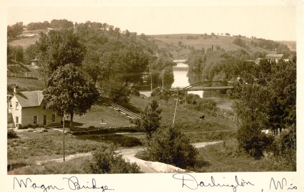 Elevated view of wagon bridge in Darlington, with a river on the right, a cow in a field, and a building on the left.
