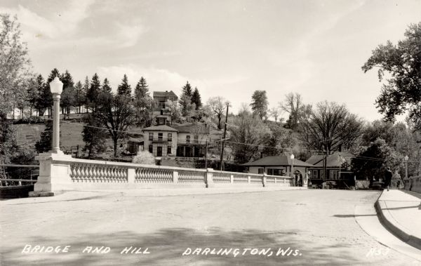 View toward bridge and hill. The bridge has lampposts. Buildings are on a hill in the background. Caption reads: "Bridge and Hill, Darlington, Wis."