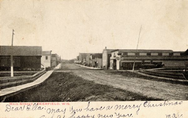 View down side of unpaved Main Street in Deerfield, with a wooden sidewalk on the left. Caption reads: "Main Street, Deerfield, Wis." Handwritten note: "Dear Ella: May you have a Merry Christmas and a happy New Year. M.A.E."