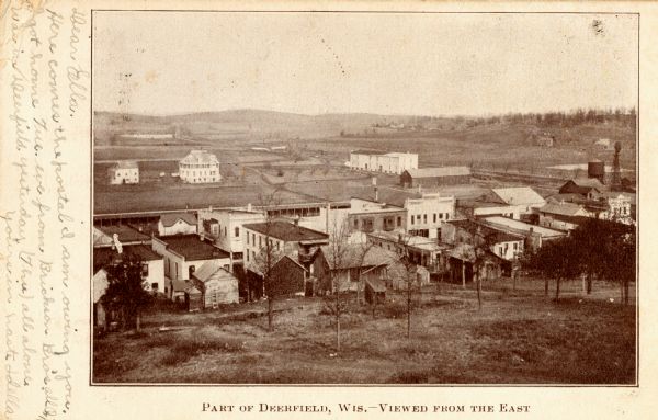 Elevated view of the town. Caption reads: "Part of Deerfield, Wis. -- viewed from the East". Handwritten note: "Dear Ella, Here comes the postal I am owing you. I got home Tue. eve from Brickson Bros. all in. Was in Deerfield yesterday (Thu) all alone. Yours in haste, Idella."