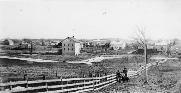 View of buildings in Delafield, with two men and a dog standing near a fence in the foreground.