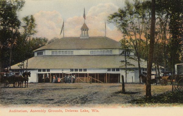 View of the Assembly Grounds Auditorium with horse-drawn carriages on the left and right, and groups of people standing just outside the building. Caption reads: "Auditorium, Assembly Grounds, Delavan Lake, Wis."