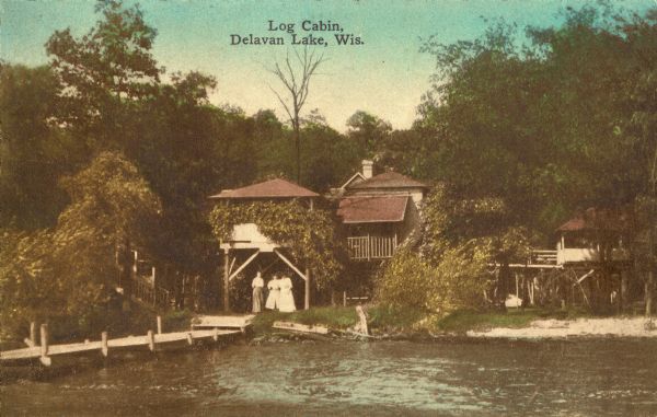 View across water towards cabins on the shore of Delavan Lake, with three women standing near the base of a pier. Caption reads: "Log Cabin, Delavan Lake, Wis."