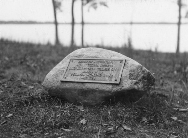 View of a rock with an engraved plate reading, "Group of prehistoric Indian burial mounds and Winnebago village site.  Marked by the Delavan Woman's Club - May 1924."
