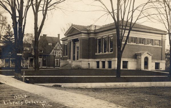 Exterior of library with unfinished landscaping. Houses are further down the street on the left. A sign attached to a tree reads: "S Fourth St." Caption reads: "Library Delavan, Wis."
