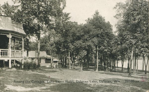 View of the lake-side of the hotel. The hotel porch and balcony is on the far left, and beyond are smaller buildings among trees. The lake is on the right. Caption reads: "Among the Cottages, Lake Lawn Hotel, Delavan Lake, Delavan, Wis."
