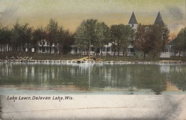 View across water towards Lake Lawn. Two turrets or towers are above the trees. The word "LAWN" is spelled out on the shorelineon the left. Caption reads: "Lake Lawn, Delavan Lake, Wis."