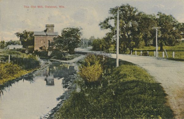 Distant view of the mill, with a stream leading to it, and a road paralleling the stream on the right. Caption reads: "The Old Mill, Delavan, Wis."